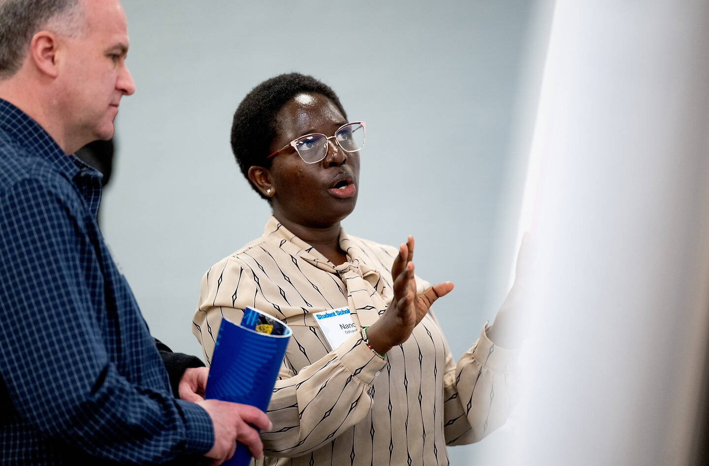 A person in a patterned shirt and glasses gestures while speaking to another individual holding a blue folder, in what appears to be a professional or educational setting.